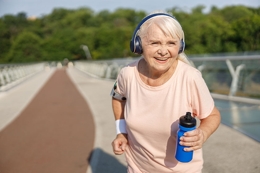 Positive senior lady with headphones runs along bridge in summer