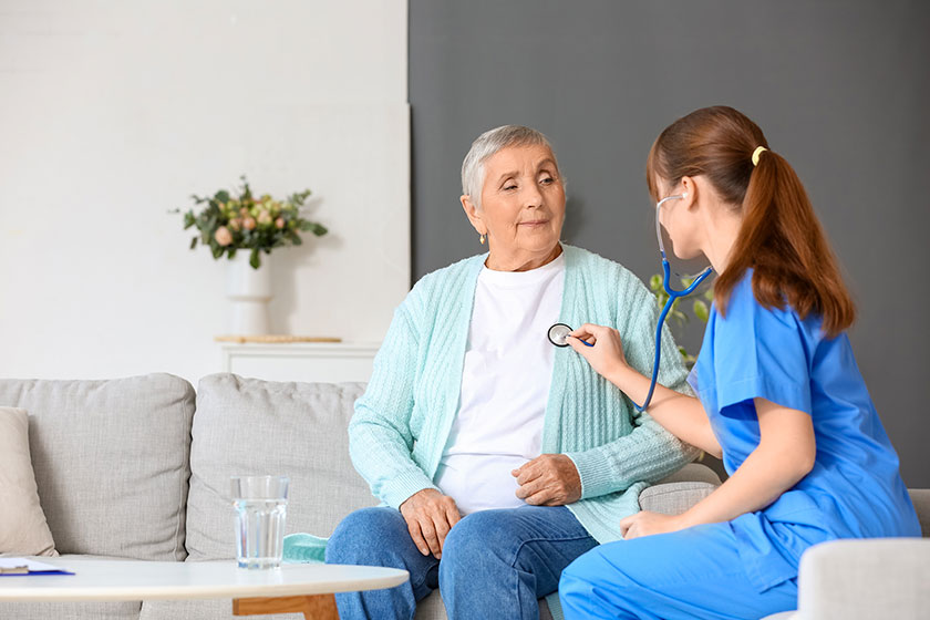 Young caregiver hearing senior woman with stethoscope home